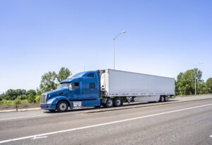 Big rig blue bonnet semi truck with high cab and long refrigerator semi trailer transporting commercial cargo load driving on the wide highway road with bridge and concrete safety fence on the side